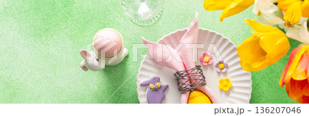 A beautifully arranged Easter table setting showcases a decorative egg nestled in a pink napkin, surrounded by a glass of water and fresh flowers, perfect for holiday gatherings, banner 136207046