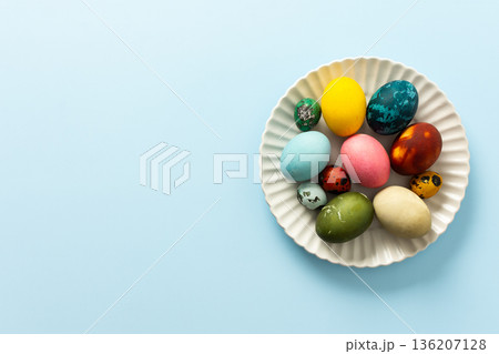 A variety of decorated Easter eggs in vibrant colors are creatively arranged on a white plate set against a soft blue background, symbolizing spring festivities and joy A variety of decorated Easter eggs in vibrant colors are creatively arranged on a white plate set against a soft blue background, symbolizing spring festivities and joy 136207128