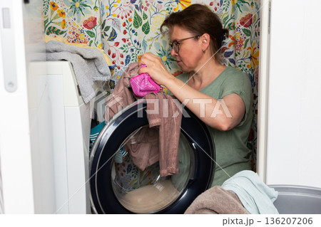 Woman stands in bathroom putting clothes into washing machine while surrounded by towels and laundry, carrying out daily household task with focus and care 136207206