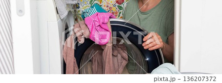 Woman stands in bathroom putting clothes into washing machine while surrounded by towels and laundry, carrying out daily household task with focus and care, banner 136207372