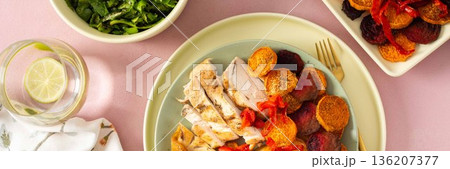 Baked potato is served with chicken breast and roasted carrots, peppers, and beets on a plate. A light meal is arranged on a simple table setting, banner 136207377