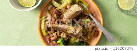 Breakfast includes baked hake placed on boiled beans with broccoli and peppers. A fresh tomato salad and lime water are nearby on a green table, banner 136207379