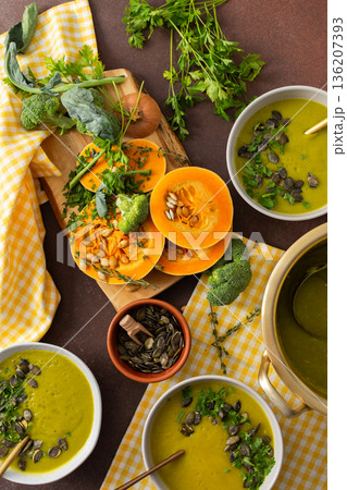 A table displays bowls of pumpkin soup with broccoli topped with seeds and herbs. Nearby, fresh ingredients and a pot hold more soup, creating a healthy meal setting 136207393
