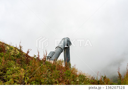 Avalanche control cannon installed on mountain slope, used for controlled snow release . Industrial safety equipment integrated into natural alpine landscape under cloudy and foggy weather conditions 136207528