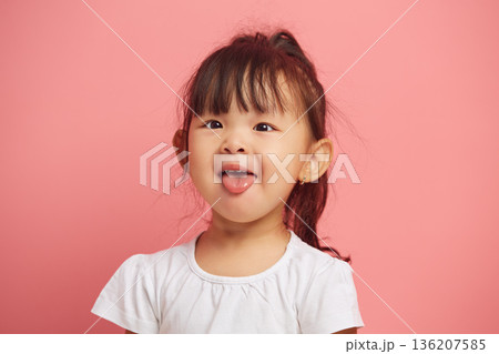 Cheerful mischievous little asian appearance girl shows her tongue to the camera, smiling cheerfully while standing on a pink polished background. Joyful child headshot portrait. 136207585