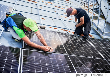 Workers building solar panel system on metal rooftop of house. Two men installers installing photovoltaic solar module outdoors. Alternative, green and renewable energy generation concept. Workers building solar panel system on metal rooftop of house. Two men installers installing photovoltaic solar module outdoors. Alternative, green and renewable energy generation concept. 136208128