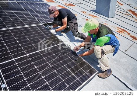Workers building solar panel system on metal rooftop of house. Two men installers installing photovoltaic solar module outdoors. Alternative, green and renewable energy generation concept. Workers building solar panel system on metal rooftop of house. Two men installers installing photovoltaic solar module outdoors. Alternative, green and renewable energy generation concept. 136208129