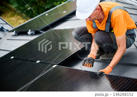 Worker building photovoltaic solar panel system on rooftop of house. Man engineer in helmets and gloves installing solar module with help of hex key outdoors. Alternative energy generation concept. Worker building photovoltaic solar panel system on rooftop of house. Man engineer in helmets and gloves installing solar module with help of hex key outdoors. Alternative energy generation concept. 136208152