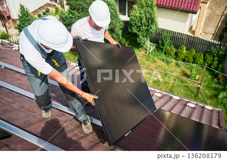 Workers building solar panel system on rooftop of house. Two men installers in helmets installing photovoltaic solar module outdoors. Alternative, green and renewable energy generation concept. Workers building solar panel system on rooftop of house. Two men installers in helmets installing photovoltaic solar module outdoors. Alternative, green and renewable energy generation concept. 136208179