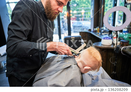 Barber using comb and shaver to cut hair. Professional hairdresser shaving little kid's nape. 136208209