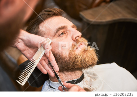 Close-up picture of handsome man getting his beard done. Male customer relaxing while professional hairdresser cutting his beard. Experienced barber using comb and scissors. 136208235