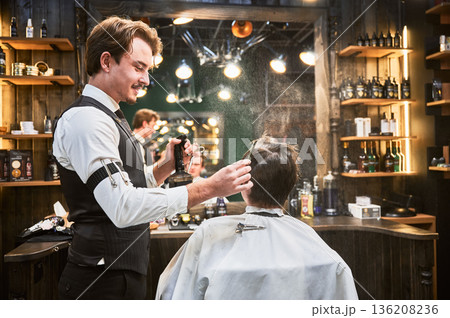 Handsome barber spraying water over head. Professional hairdresser preparing hair for cutting. Young client getting stylish hairstyle. 136208236