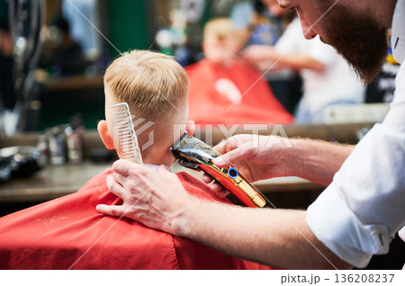 Barber using comb and shaver to cut hair. Professional hairdresser shaving little kid's nape. Close up. Barber using comb and shaver to cut hair. Professional hairdresser shaving little kid's nape. Close up. 136208237