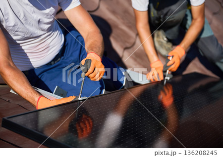 Workers building photovoltaic solar panel system on rooftop of house. Close up of men technicians in helmets and gloves installing solar module with help of hex key outdoors. Renewable energy. Workers building photovoltaic solar panel system on rooftop of house. Close up of men technicians in helmets and gloves installing solar module with help of hex key outdoors. Renewable energy. 136208245