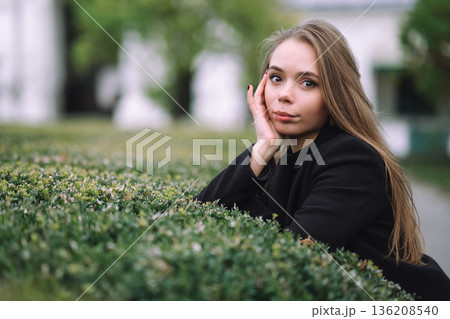 Thoughtful Young Woman Portrait Outdoors Leaning on Green Hedge in Urban Park with Soft Natural Light and Calm Elegant Mood 136208540