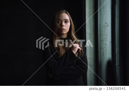 Moody Young Woman Portrait Standing Near Window Indoors with Natural Side Light and Dark Minimal Background Creating Cinematic Atmosphere Moody Young Woman Portrait Standing Near Window Indoors with Natural Side Light and Dark Minimal Background Creating Cinematic Atmosphere 136208541