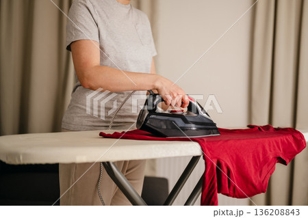 Woman ironing a long sleeve clothing at home. Daily chores. Laundry day. Neutral background. Home appliances Woman ironing a long sleeve clothing at home. Daily chores. Laundry day. Neutral background. Home appliances 136208843