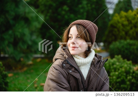 Young woman wearing warm jacket and hat sitting in a green city park. 136209513