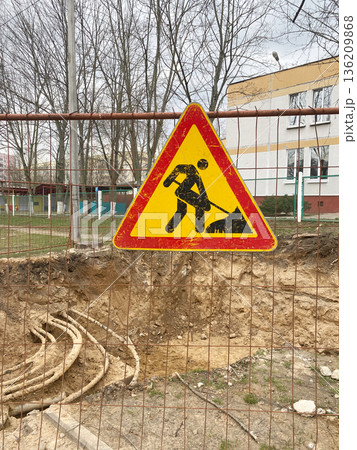 Caution sign indicating construction work ahead, featuring a worker with a shovel, surrounded by a fenced area with exposed earth and trees in the background, emphasizing safety awareness Caution sign indicating construction work ahead, featuring a worker with a shovel, surrounded by a fenced area with exposed earth and trees in the background, emphasizing safety awareness 136209868