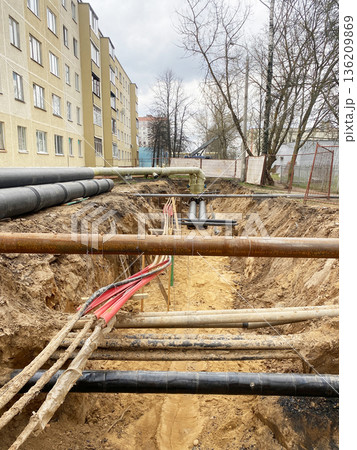 Construction site showing deep trench with various pipes and cables, surrounded by buildings and trees, illustrating urban infrastructure development and maintenance Construction site showing deep trench with various pipes and cables, surrounded by buildings and trees, illustrating urban infrastructure development and maintenance 136209869