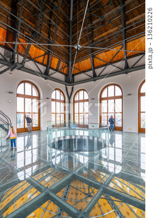 Visitors explore the tower of the National Museum in Prague. The glass floor reveals intricate designs below. Large windows provide views of the city. People engage with the space. 136210226