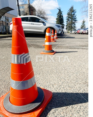 Bright orange traffic cones lined up on a paved parking lot, with a white vehicle parked nearby, showcasing safety measures and urban infrastructure in a sunny environment Bright orange traffic cones lined up on a paved parking lot, with a white vehicle parked nearby, showcasing safety measures and urban infrastructure in a sunny environment 136211265