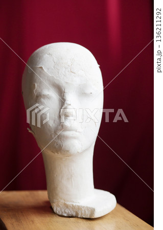 White plaster mannequin head displayed on wooden surface against a rich red backdrop, showcasing artistic design and texture, ideal for creative projects and visual storytelling White plaster mannequin head displayed on wooden surface against a rich red backdrop, showcasing artistic design and texture, ideal for creative projects and visual storytelling 136211292