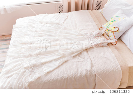Elegant wedding dress delicately laid on a soft bed, surrounded by light fabrics and a decorative pillow, creating a serene atmosphere for bridal preparations Elegant wedding dress delicately laid on a soft bed, surrounded by light fabrics and a decorative pillow, creating a serene atmosphere for bridal preparations 136211326