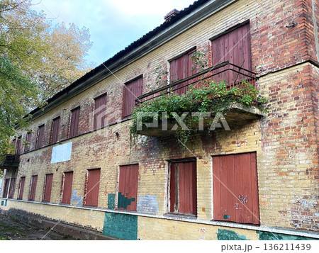 Abandoned building with weathered brick walls, boarded windows, and overgrown balcony, showcasing nature reclaiming urban space in a serene environment 136211439
