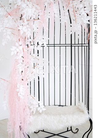 Elegant decorative birdcage adorned with white leaves and pink accents, featuring a plush fur seat, creating a serene and inviting atmosphere for interior design 136211443