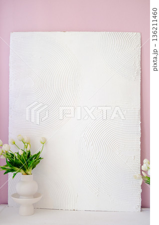 Textured white wall art with circular patterns, complemented by a minimalist vase of fresh flowers, creating a serene and stylish interior decor atmosphere. Copy space 136211460