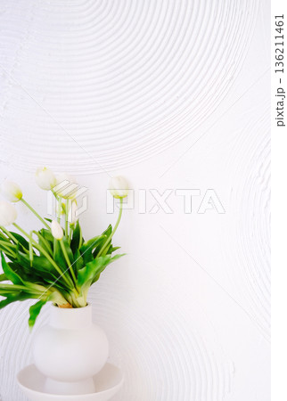 White vase filled with fresh tulips sits against a textured white wall, creating a serene and elegant atmosphere, perfect for home decor or floral arrangements. Copy space 136211461