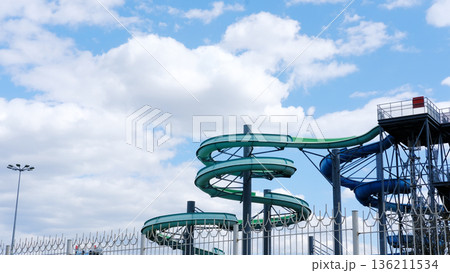 Empty water park featuring colorful water slides against a bright blue sky with fluffy clouds, creating a serene and tranquil atmosphere with ample copy space for design Empty water park featuring colorful water slides against a bright blue sky with fluffy clouds, creating a serene and tranquil atmosphere with ample copy space for design 136211534
