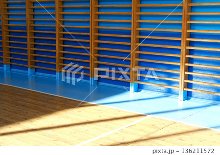 Sunlight streaming through wooden slats illuminates a vibrant blue wall and polished wooden floor, creating a warm and inviting atmosphere in a spacious interior setting Sunlight streaming through wooden slats illuminates a vibrant blue wall and polished wooden floor, creating a warm and inviting atmosphere in a spacious interior setting 136211572