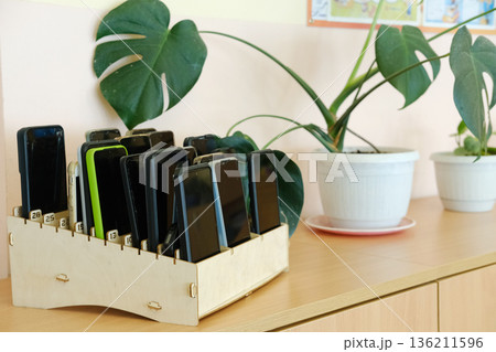 Schoolchildren's mobile phones neatly organized in a wooden box on a classroom shelf, emphasizing the ban on devices during educational activities and promoting focus on learning 136211596