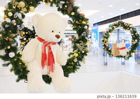 Plush teddy bear wearing red scarf hangs from festive wreath adorned with ornaments, creating a cheerful holiday atmosphere in a bustling shopping center Plush teddy bear wearing red scarf hangs from festive wreath adorned with ornaments, creating a cheerful holiday atmosphere in a bustling shopping center 136211639