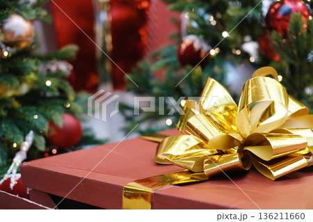 Festive gift box adorned with a shiny gold ribbon and bow, surrounded by decorated Christmas trees and sparkling ornaments, creating a joyful holiday atmosphere 136211660