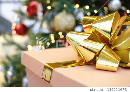 Gift box adorned with a shiny gold ribbon, placed in front of a beautifully decorated Christmas tree, creating a festive and joyful holiday atmosphere Gift box adorned with a shiny gold ribbon, placed in front of a beautifully decorated Christmas tree, creating a festive and joyful holiday atmosphere 136211678