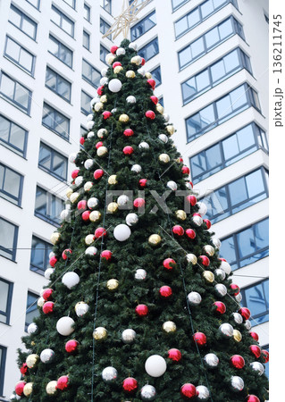Christmas tree adorned with colorful ornaments, standing tall against a modern building, showcasing festive spirit and holiday cheer in an urban environment Christmas tree adorned with colorful ornaments, standing tall against a modern building, showcasing festive spirit and holiday cheer in an urban environment 136211745