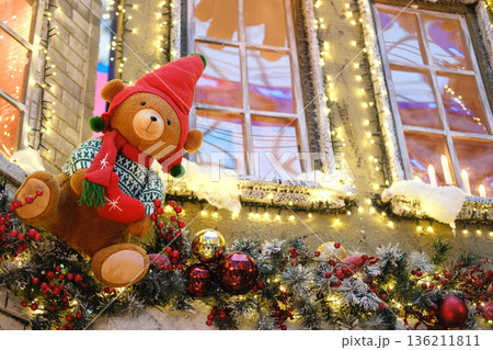 Plush teddy bear wearing a festive red hat and scarf is perched on a beautifully decorated garland with sparkling lights in a vibrant shopping center during the holiday season Plush teddy bear wearing a festive red hat and scarf is perched on a beautifully decorated garland with sparkling lights in a vibrant shopping center during the holiday season 136211811