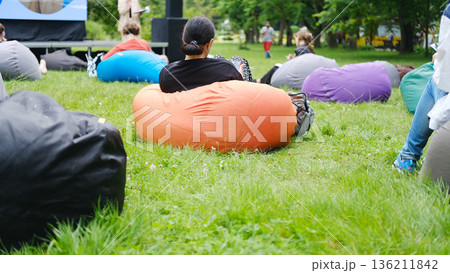 Group of individuals seated on colorful beanbags in a lush green park, attentively listening to an outdoor lecture, creating a relaxed learning atmosphere with copy space 136211842