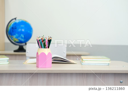 Colorful pencils in pink holder on wooden desk with open book and stacked notebooks, globe in background, classroom setting with light walls and furniture Colorful pencils in pink holder on wooden desk with open book and stacked notebooks, globe in background, classroom setting with light walls and furniture 136211860
