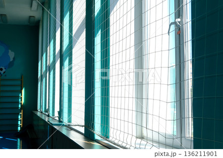 Brightly lit school gym featuring large windows adorned with protective netting, casting soft shadows on the floor, creating a safe and inviting atmosphere for students and activities Brightly lit school gym featuring large windows adorned with protective netting, casting soft shadows on the floor, creating a safe and inviting atmosphere for students and activities 136211901