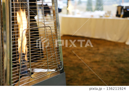Outdoor patio heater with flame visible in foreground, blurred table with white tablecloth and dishes in background, grass surface beneath, evening setting with soft lighting Outdoor patio heater with flame visible in foreground, blurred table with white tablecloth and dishes in background, grass surface beneath, evening setting with soft lighting 136211924