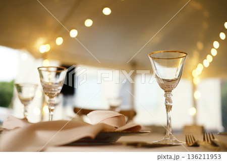 Elegant glassware on table with white napkin and silver cutlery under tent with string lights in background during evening event with soft lighting Elegant glassware on table with white napkin and silver cutlery under tent with string lights in background during evening event with soft lighting 136211953