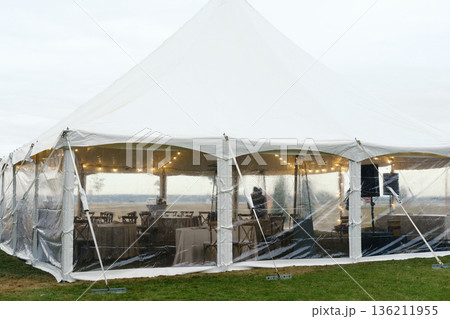 Outdoor celebration tent with transparent sides, elegantly decorated interior, providing shelter from weather elements, creating a festive atmosphere for gatherings and events Outdoor celebration tent with transparent sides, elegantly decorated interior, providing shelter from weather elements, creating a festive atmosphere for gatherings and events 136211955