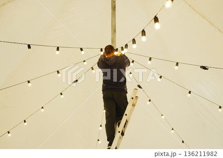 Male technician adjusting decorative string lights on a ladder inside a large tent, creating a warm ambiance for an outdoor event with illuminated atmosphere 136211982