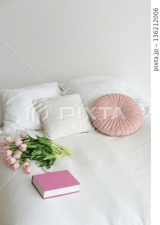 Cozy bedroom scene featuring soft pillows, a pink round cushion, fresh tulips, and a pink book on a white bed, ideal mockup with copy space for designer text Cozy bedroom scene featuring soft pillows, a pink round cushion, fresh tulips, and a pink book on a white bed, ideal mockup with copy space for designer text 136212006