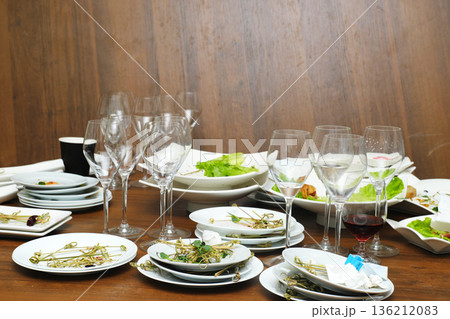 Leftover food and dirty dishes scattered on a wooden table after an event, featuring empty glasses and plates, showcasing the aftermath of a lively gathering with remnants of a meal Leftover food and dirty dishes scattered on a wooden table after an event, featuring empty glasses and plates, showcasing the aftermath of a lively gathering with remnants of a meal 136212083