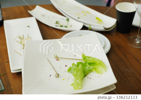 Dirty dishes piled on a wooden table after a banquet, featuring leftover food scraps, empty plates, and utensils, showcasing the aftermath of a festive reception with vibrant colors 136212113